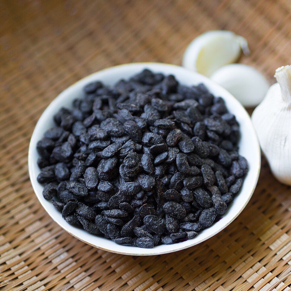 A white bowl of small black fermented soy beans beside garlic
